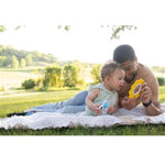 A father and toddler lying on a white blanket in a grassy park, each holding a small toy with a circular face. The father holds a yellow toy, and the toddler holds a blue-and-white toy.