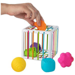 A white plastic sorting cube toy with colorful vertical bars, shown with a hand placing an orange flower-shaped piece into it. Additional flower-shaped pieces in orange, pink, and purple, as well as shell and cube pieces in green and blue, are placed outside the cube on a white background.