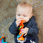 A baby wearing a dark navy hoodie holds and bites an orange, green, and blue textured teething toy while lying on a patterned carpet.
