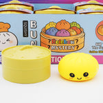 A yellow squishydumplingtoy with a cute cartoon face is shown next to its open round yellow container, with a colorful branded box in the background.