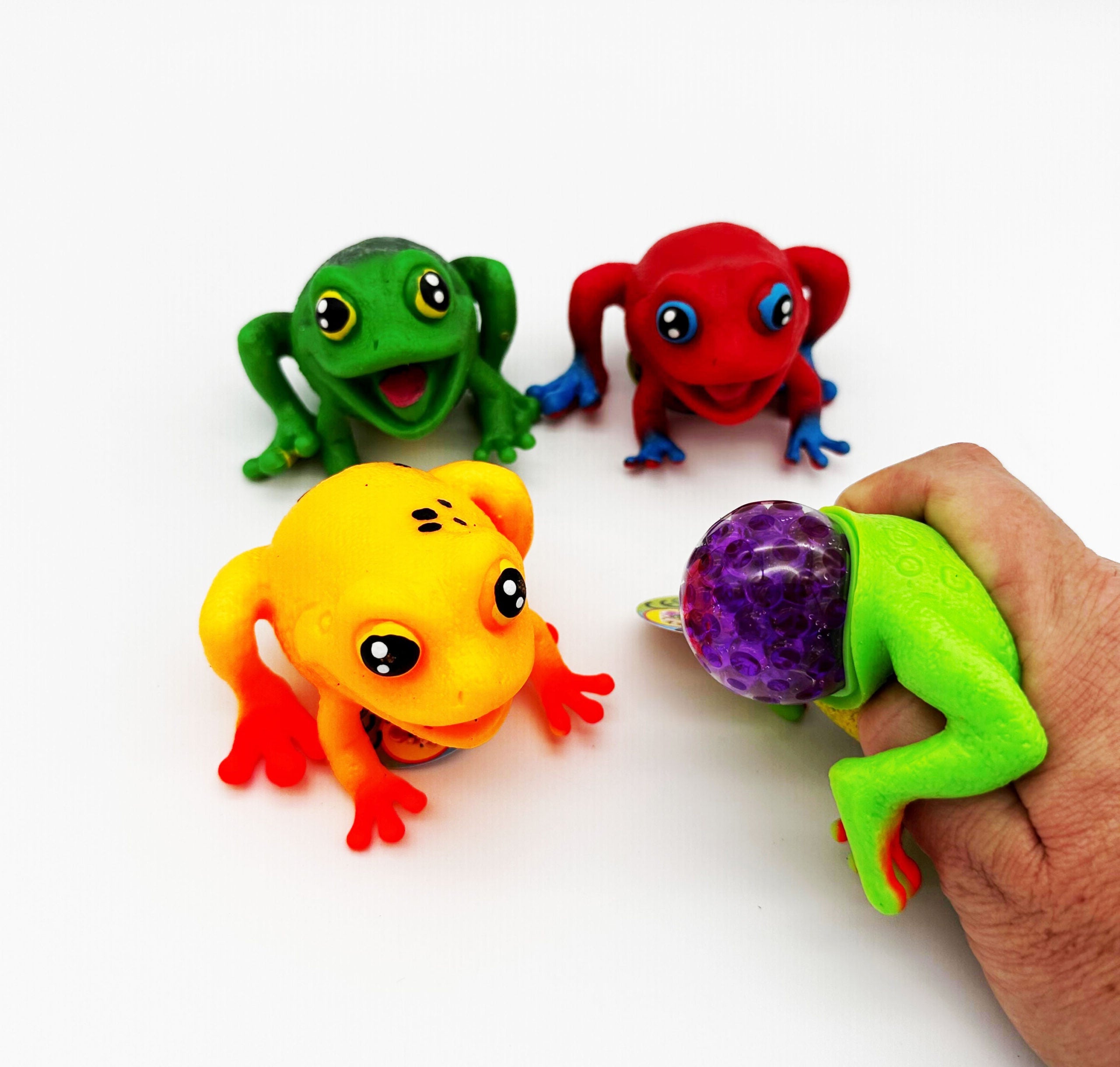 Four colorful frog-shaped squishy toys on a white background, with one being held in a human hand to show its squeeze function.